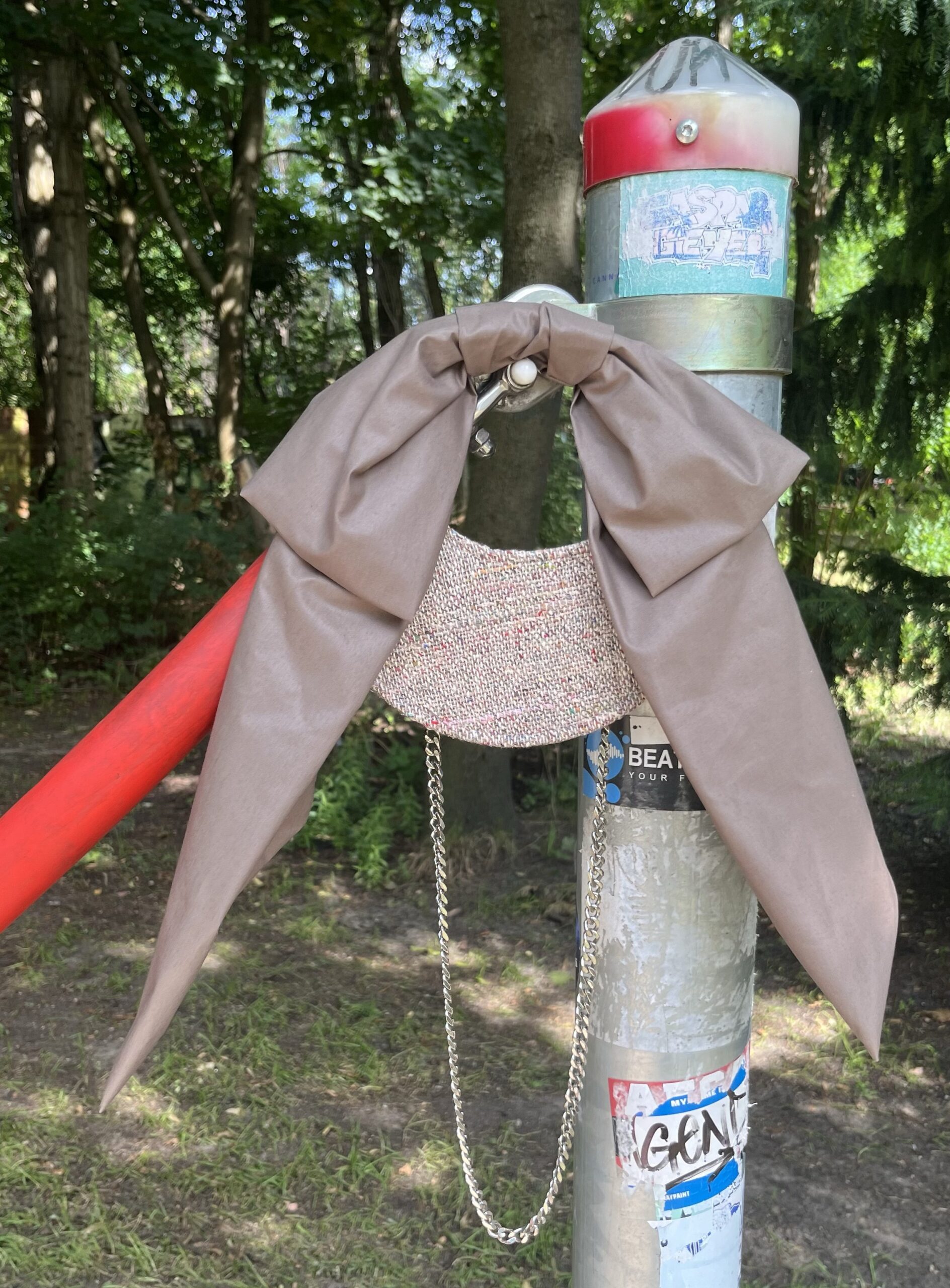 A small, structured handbag known as “The Bowby Bag S” with a taupe bow and silver chain strap hangs on a sticker-covered metal pole outdoors surrounded by trees.