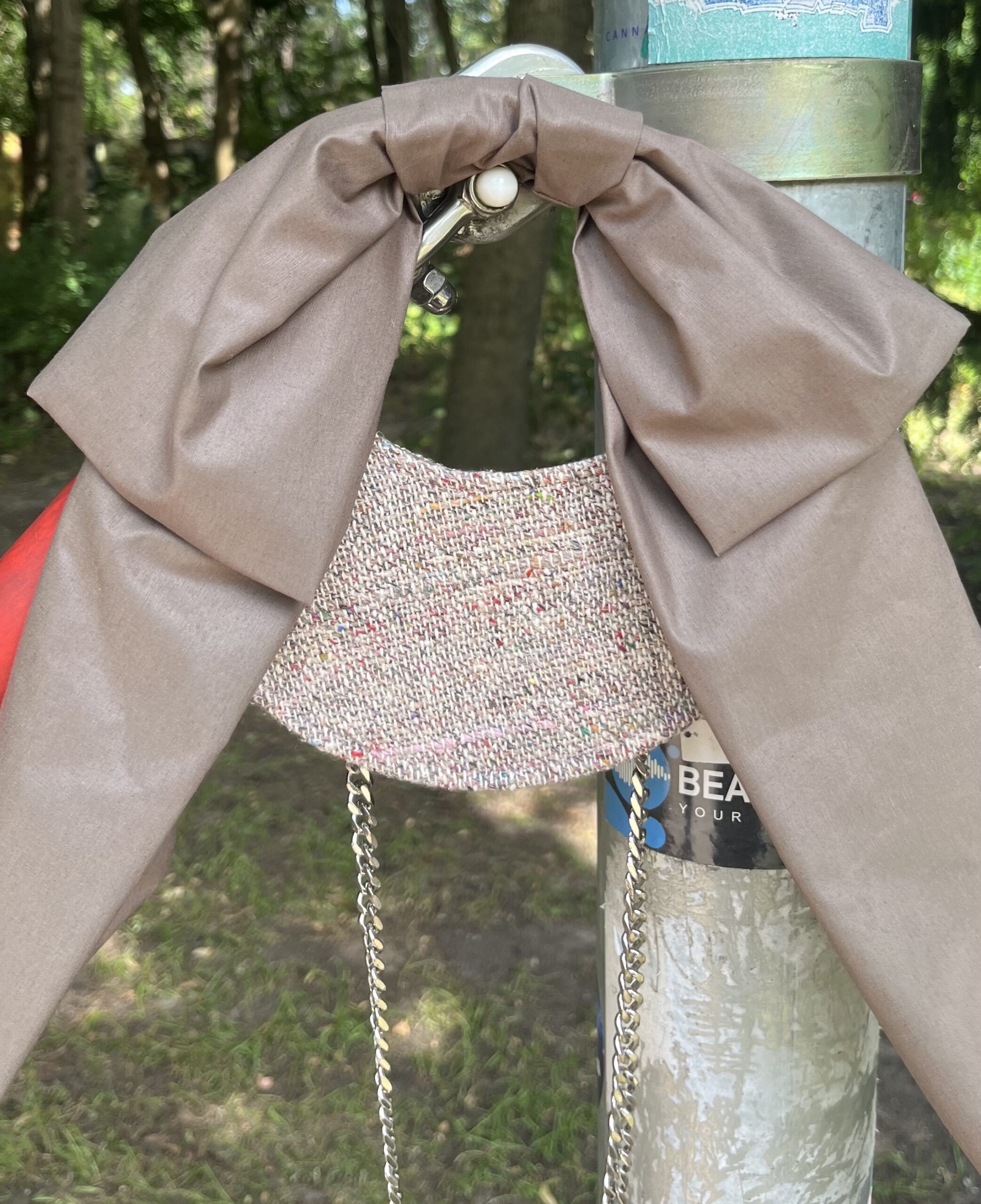 A brown fabric bow attached to a metallic ring and chain draped over a cylindrical metal pole with trees and foliage in the background.